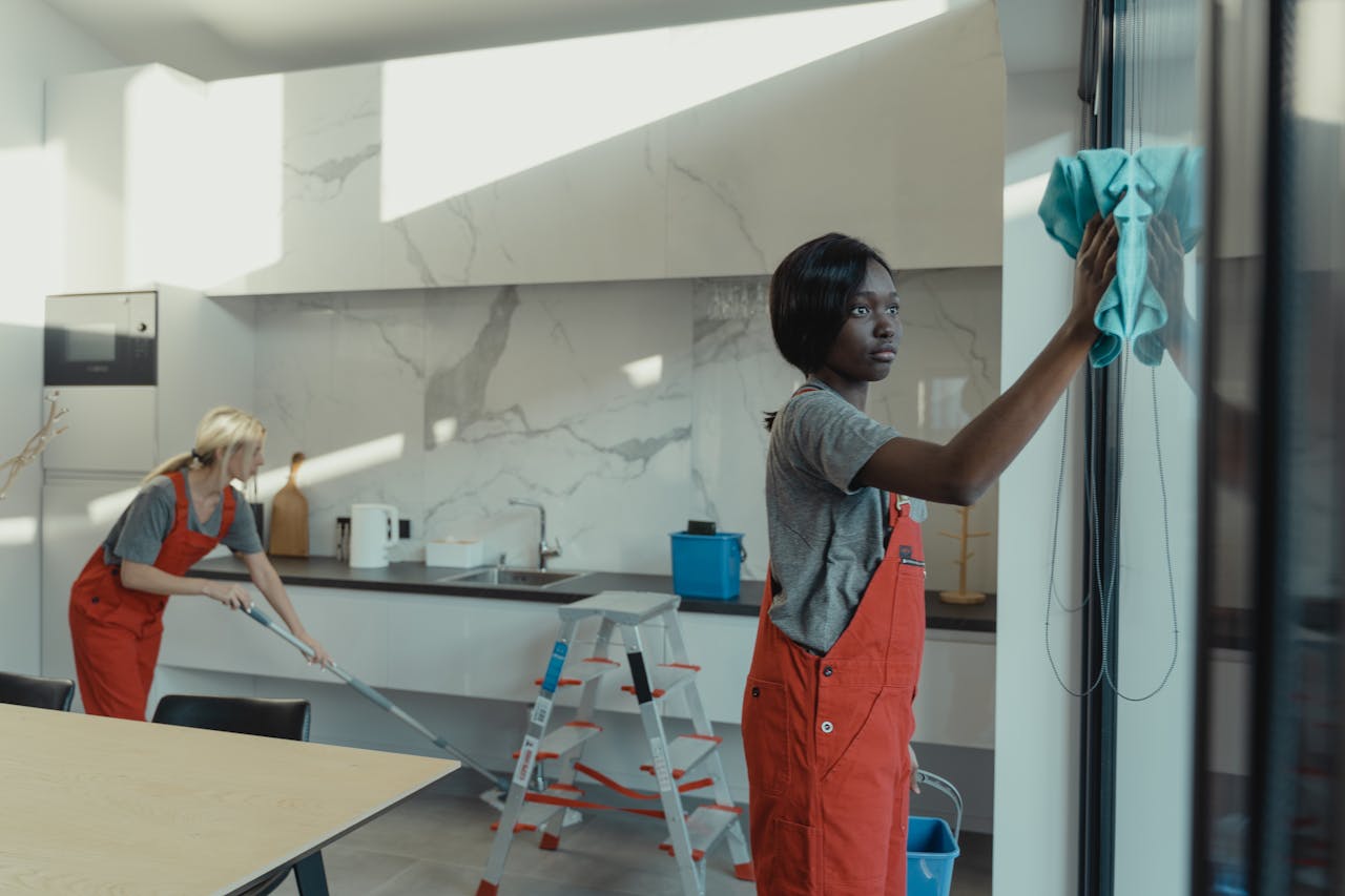 services-02 Two women cleaning a modern kitchen, wiping glass surfaces and mopping the floor.
