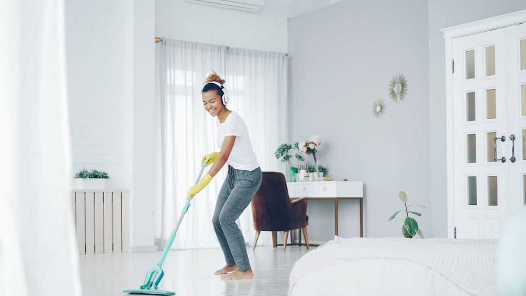 gallery-2 Cheerful African American girl in gloves is mopping floor, listening to music with wireless headphones and dancing enjoying rhythm cleaning her nice modern apartment.