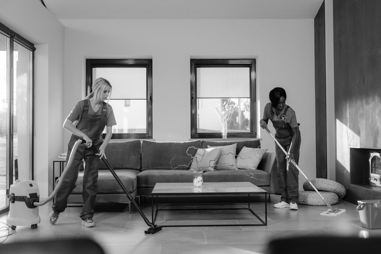 gallery-4 Two women in work clothes cleaning a modern living room using a vacuum and mop.